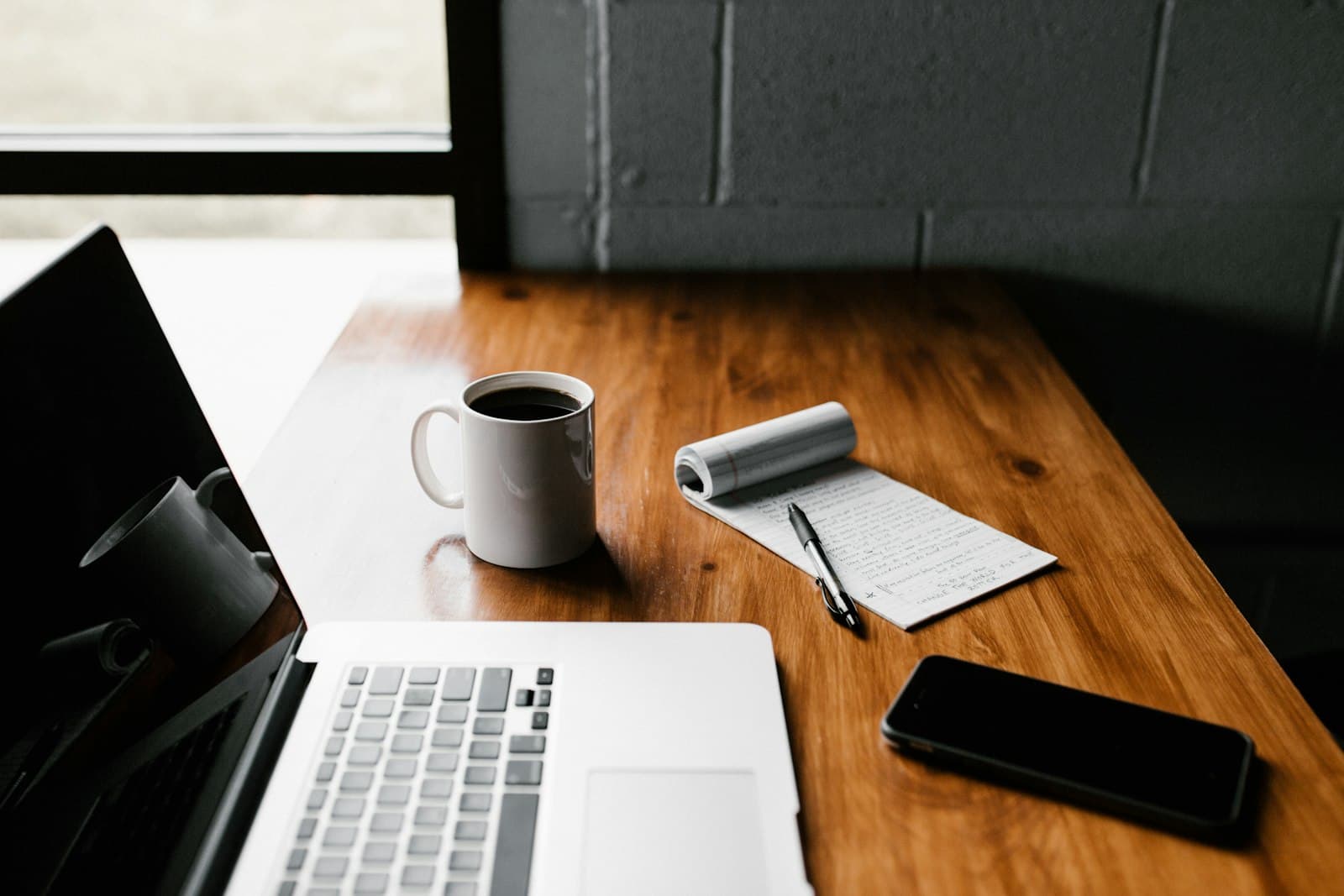A writing desk with a laptop, handwritten notes, coffee, and a phone